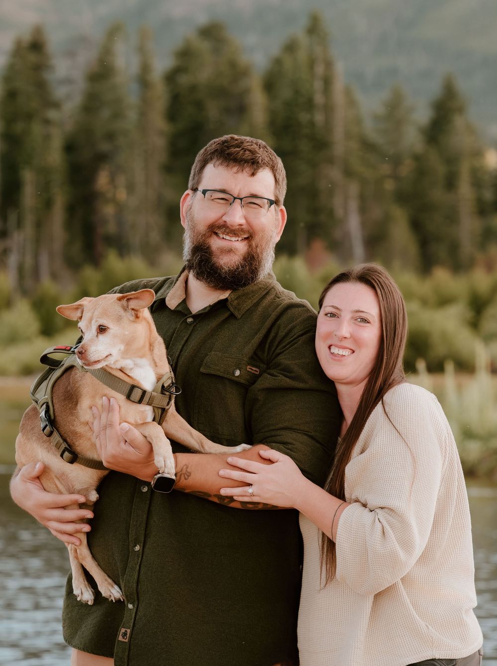 A smiling couple holds their tan dog outdoors near a lake with pine trees and mountains in the background.