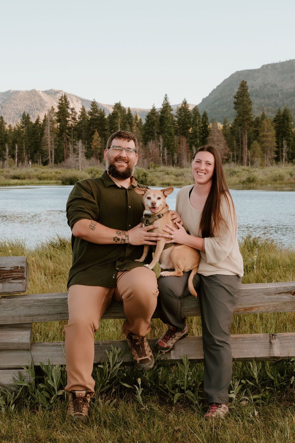 A couple smiles while holding a small dog on a wooden fence near a mountain lake surrounded by pine trees.