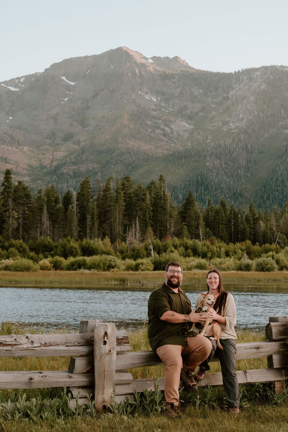 Couple smiling with dog on wooden fence, scenic mountain lake and pine forest backdrop in summer.