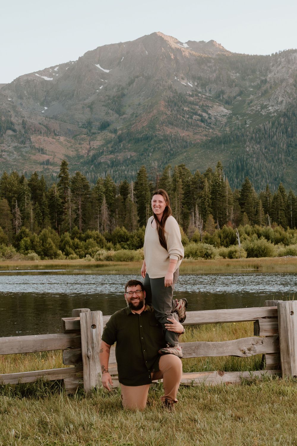 Couple posing by a wooden fence with a mountain lake and pine trees in the background during golden hour.