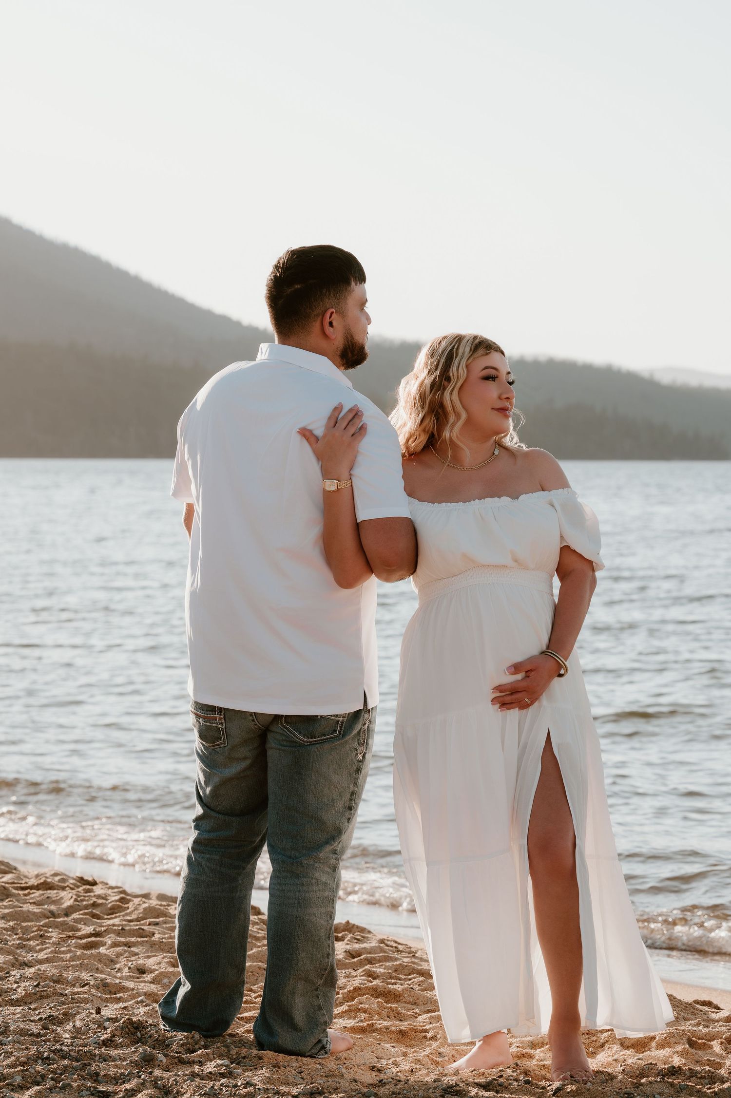 Pregnant couple in white outfits poses on sandy beach at sunset, woman in off-shoulder maternity dress cradling baby bump.