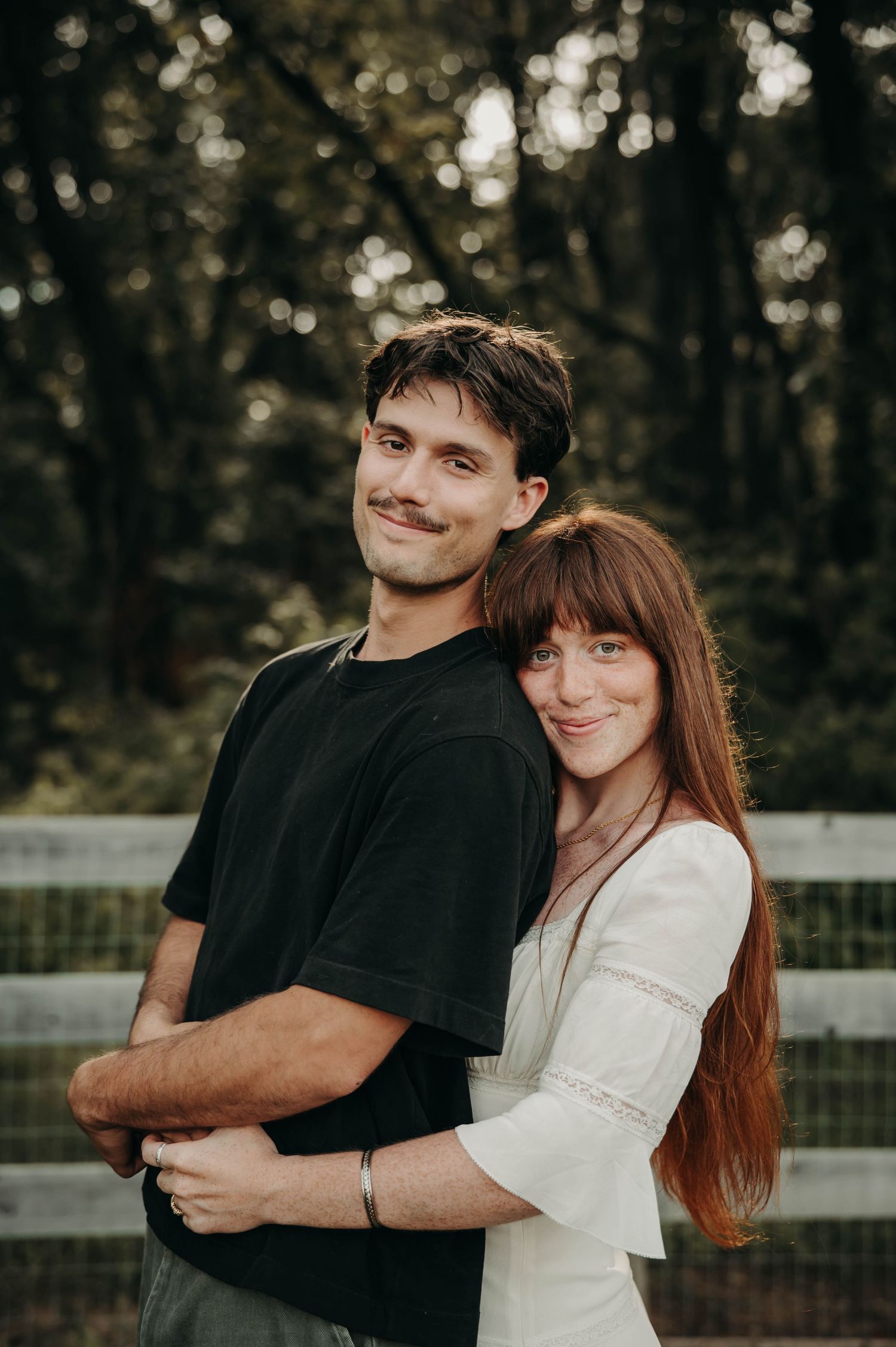 A young couple embracing outdoors near a white fence, surrounded by lush green trees in a romantic portrait.