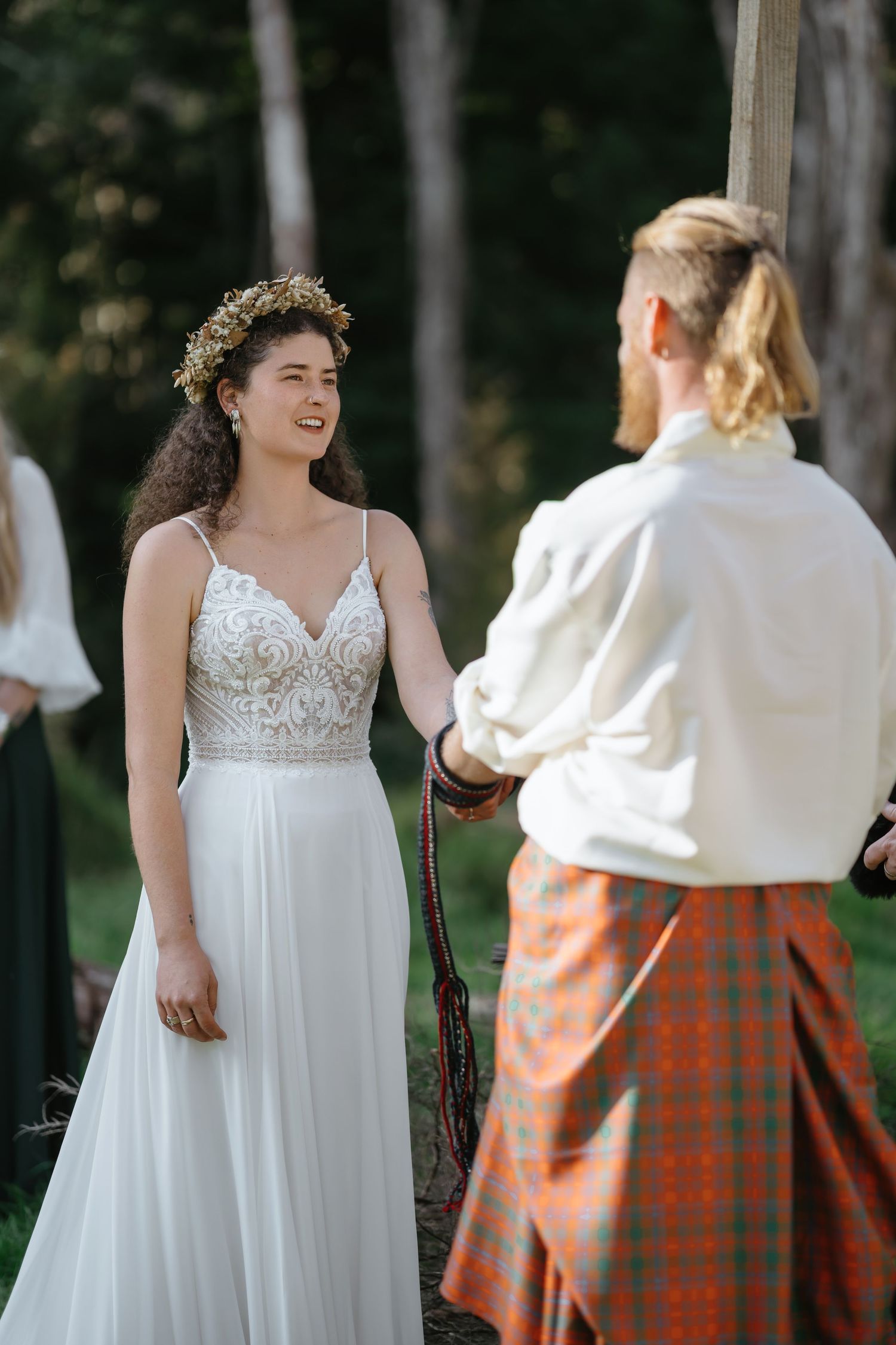 Bride in white gown with floral crown exchanges vows with partner in red tartan kilt at outdoor forest wedding.