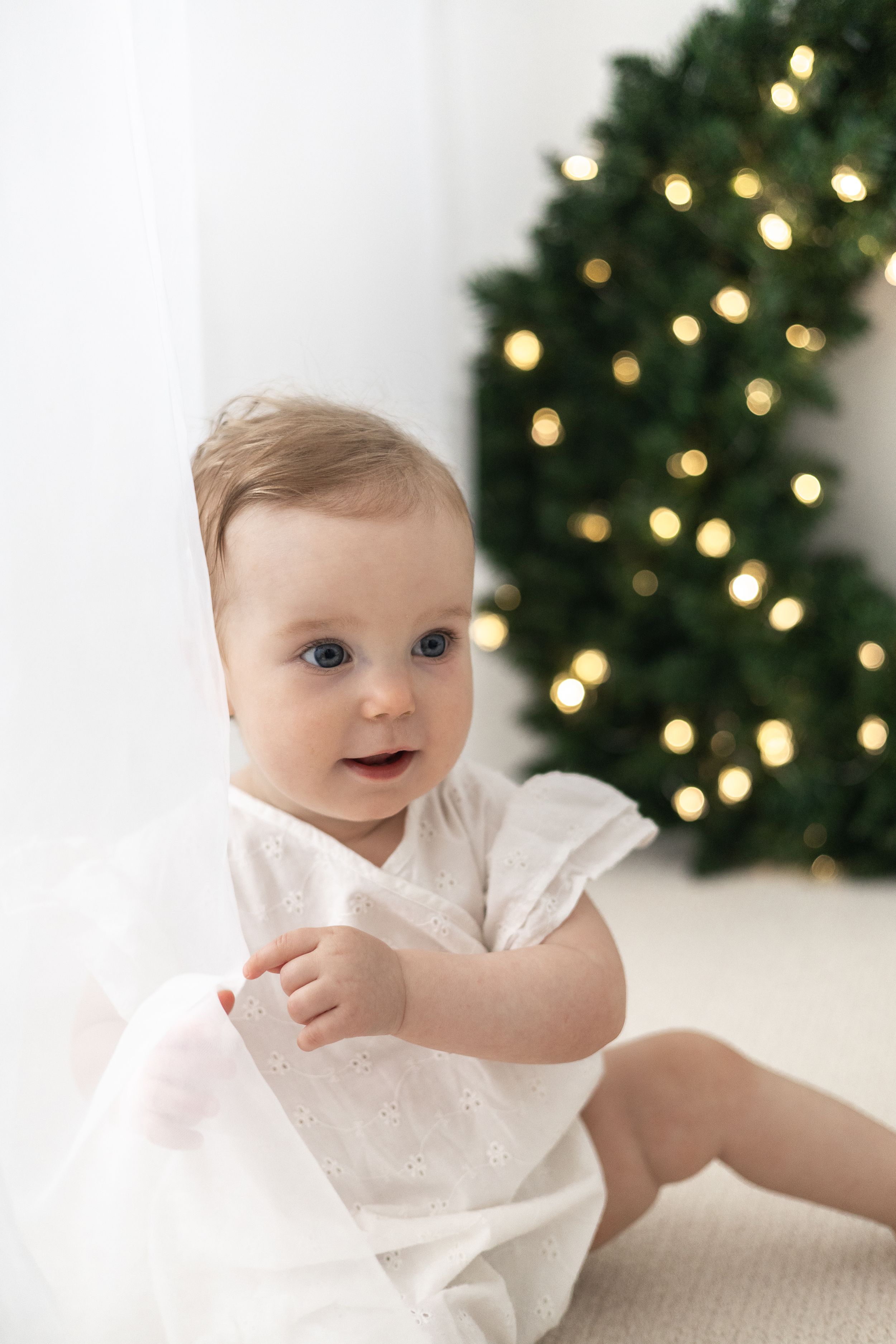 Adorable baby girl in white dress sitting on floor near decorated Christmas wreath with glowing lights Christmas mini Photoshoot Nottingham