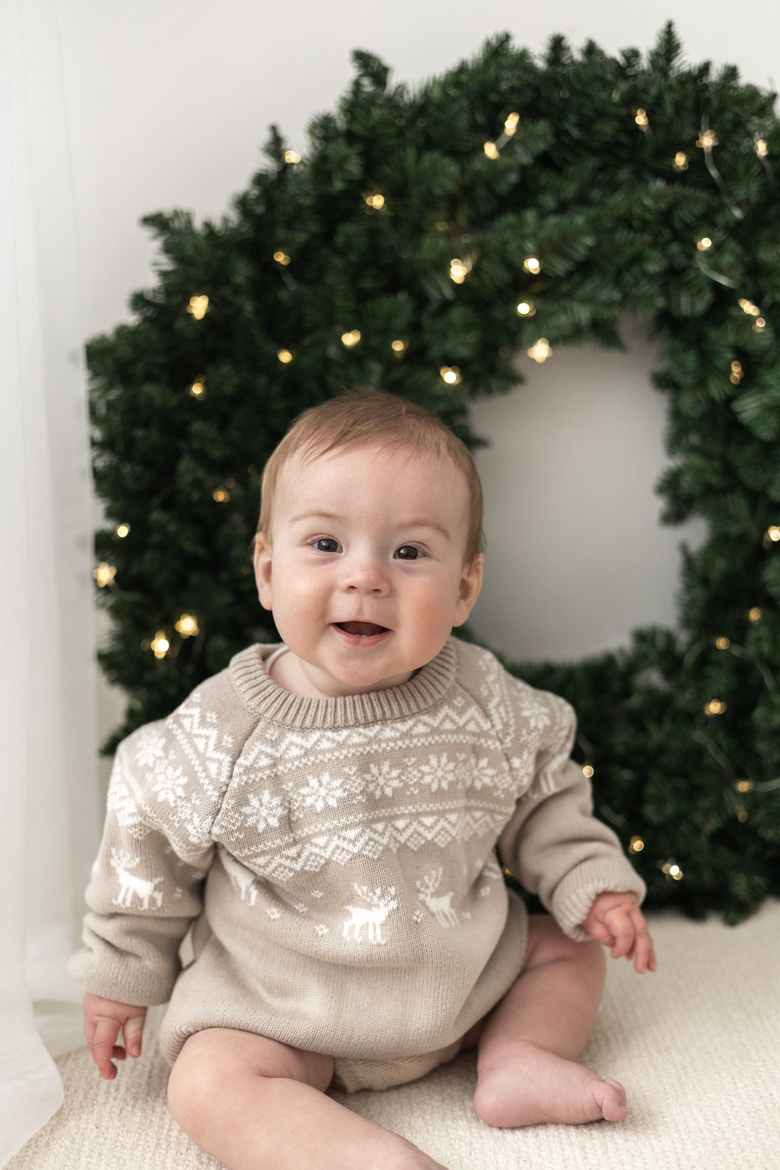 Smiling baby in Nordic reindeer sweater sits in front of a festive Christmas wreath with glowing lights Christmas mini Photoshoot Nottingham