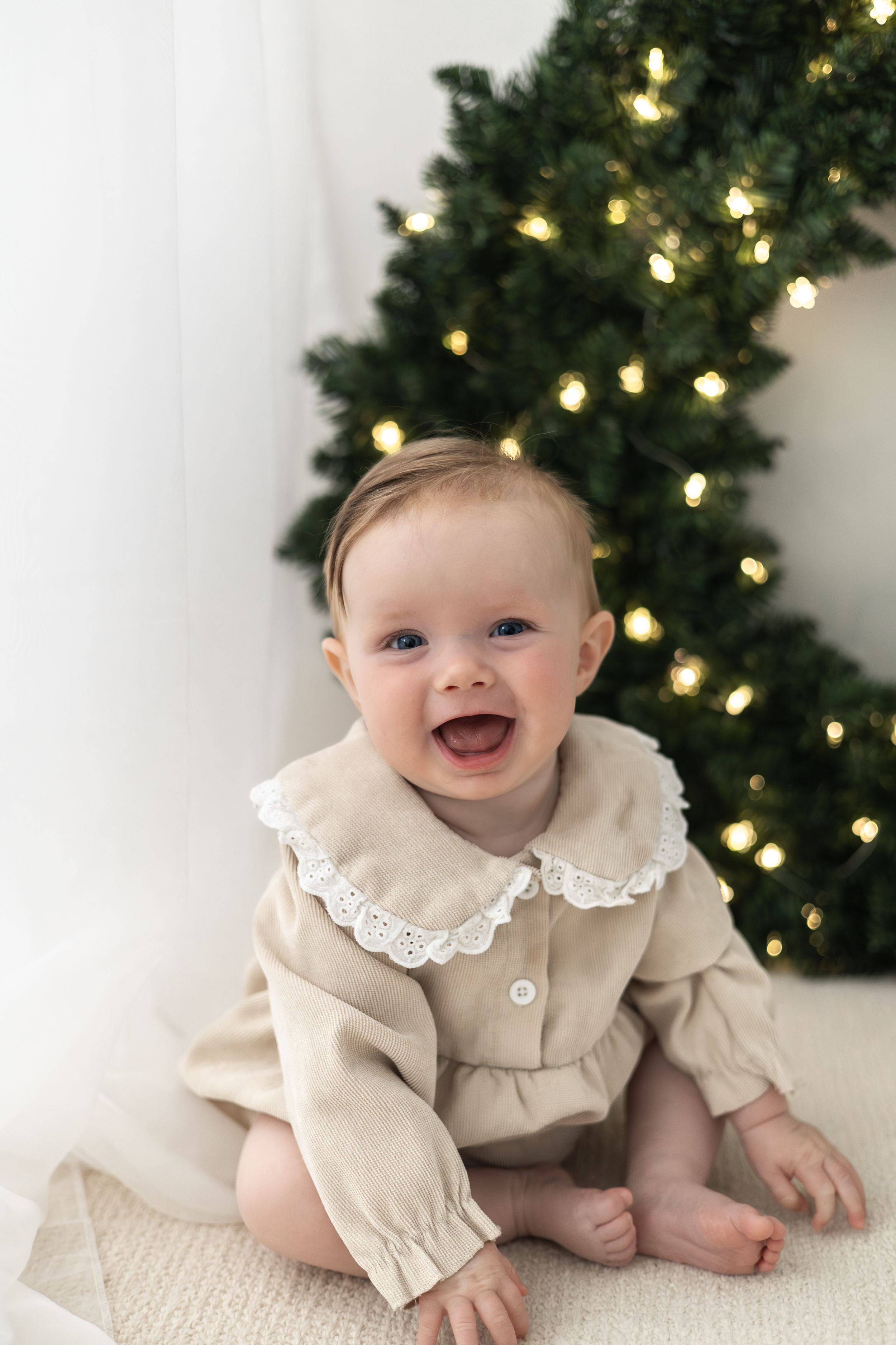 Smiling baby in beige dress sitting in front of lit Christmas wreath Christmas mini Photoshoot Nottingham