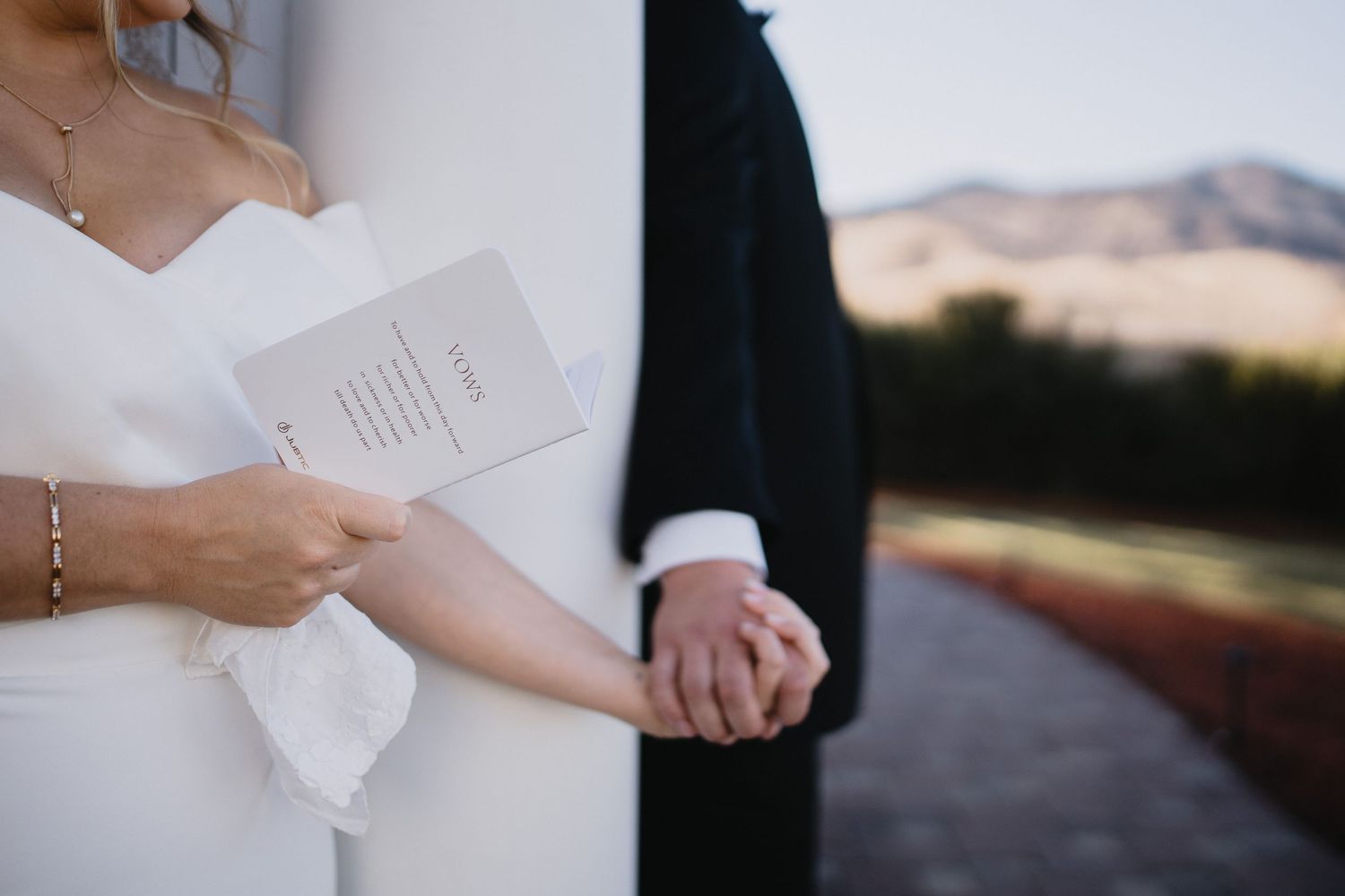 Bride in white strapless gown holds wedding program while couple holds hands outdoors with scenic hills backdrop.