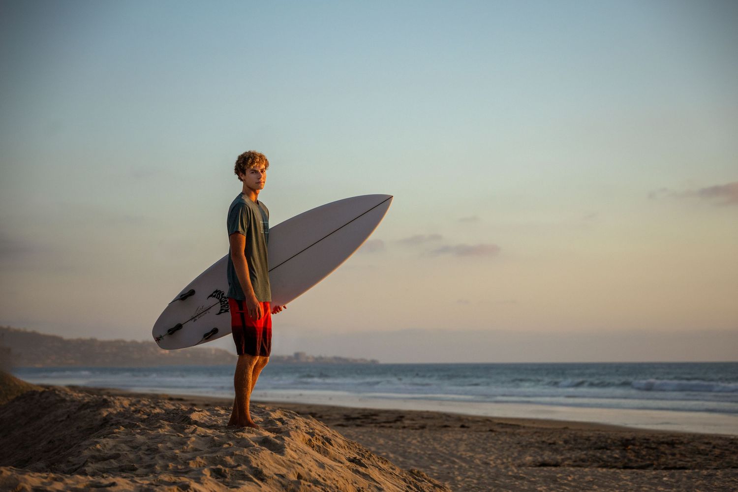 Young surfer holding a white surfboard on a sandy beach at sunset with ocean waves in the background.