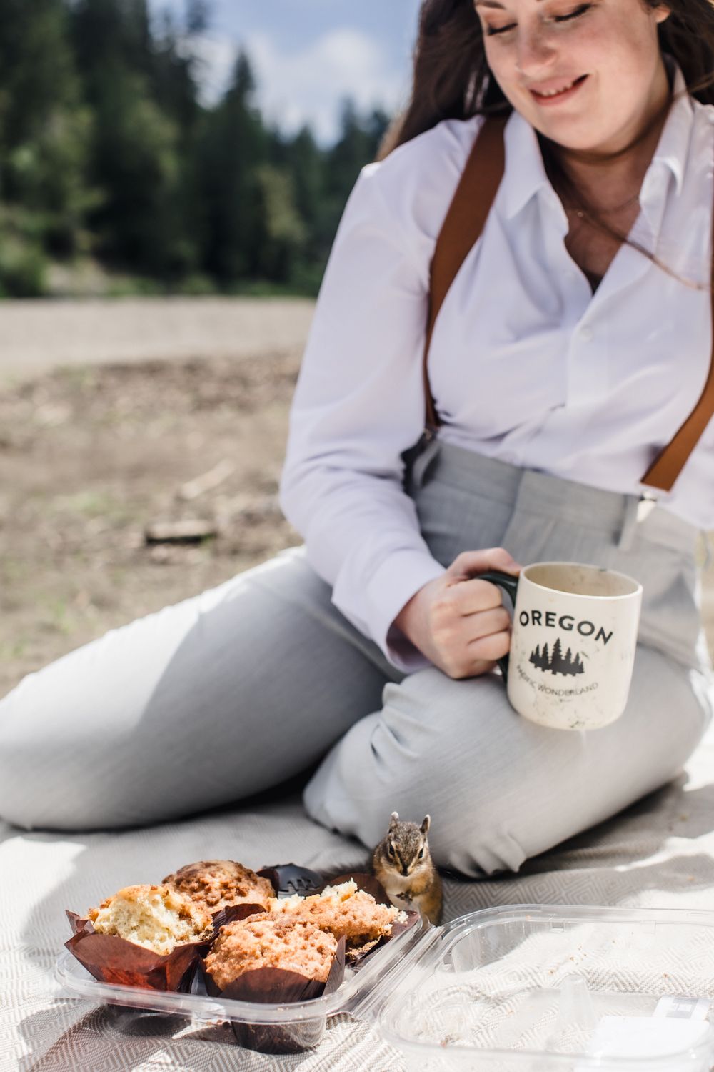 Woman sitting outdoors on a blanket holding an Oregon mug with a chipmunk eating from a food container beside her.