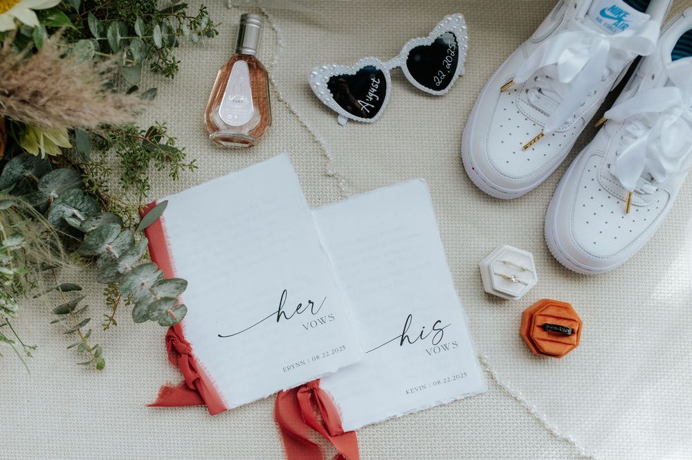 Wedding flat lay with vow books, white sneakers, heart sunglasses, rings, and floral accents on white background.