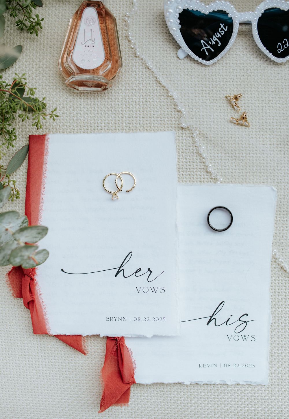 Wedding flat lay displaying his and her vow books, rings, sunglasses, and floral accents on linen.