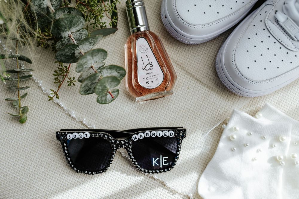 Bridal accessories flat lay with custom KE sunglasses, perfume, white sneakers, and eucalyptus greenery.