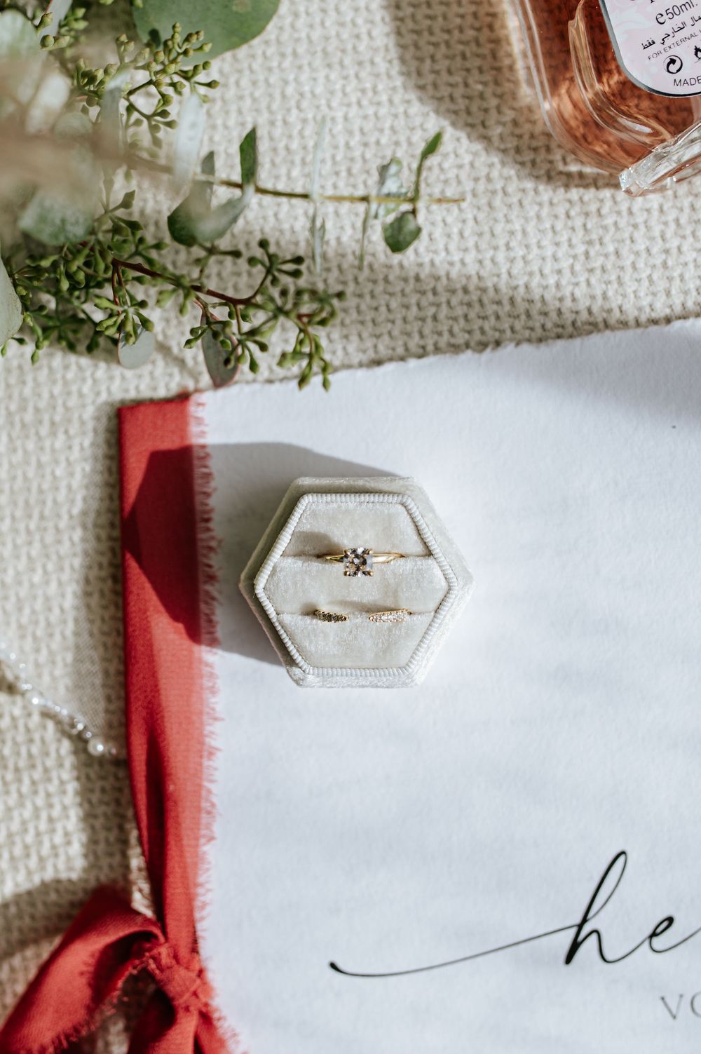 A delicate diamond engagement ring displayed in a white hexagonal velvet ring box on a wedding invitation.