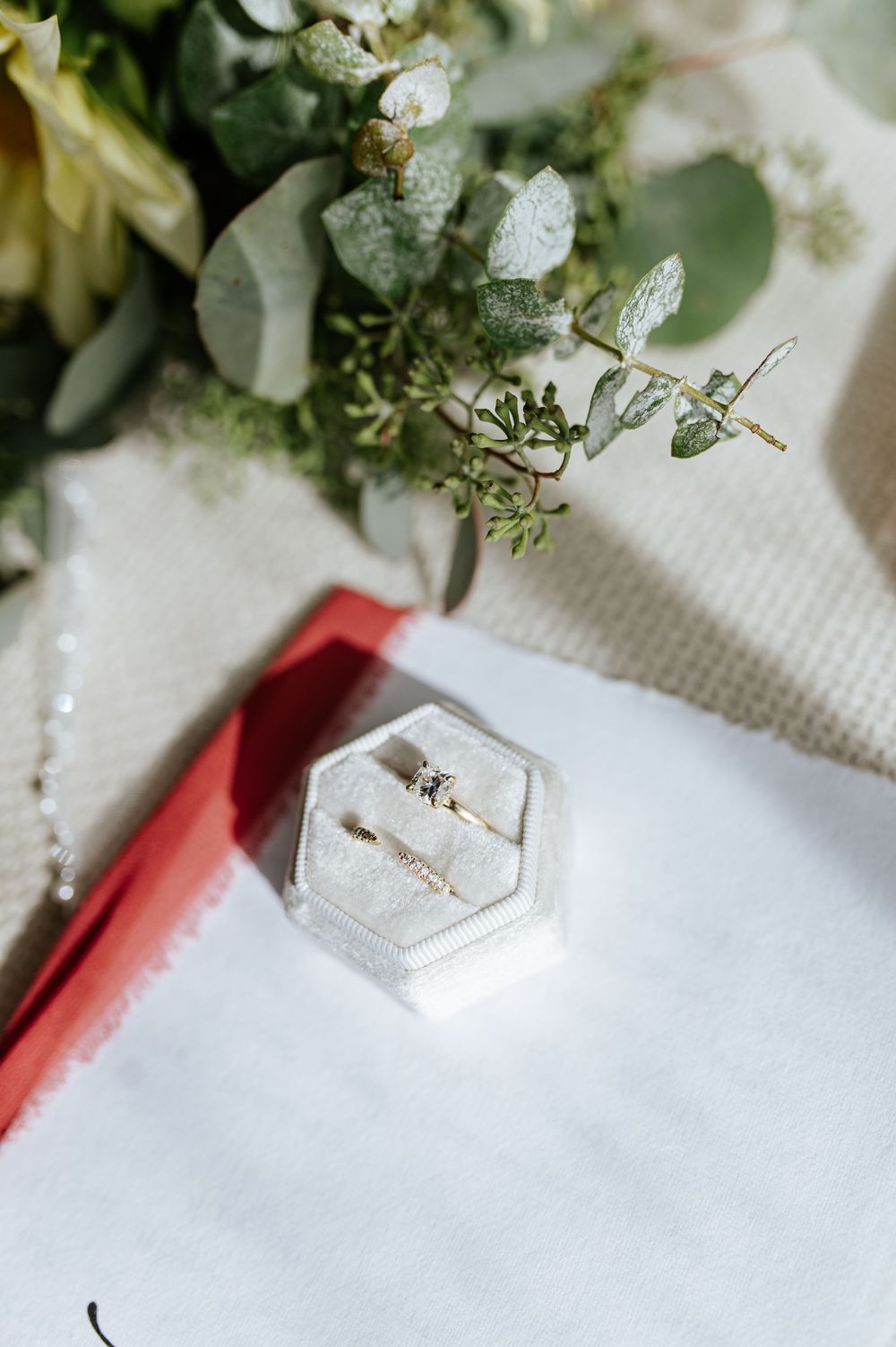 Elegant wedding rings displayed in a hexagonal velvet box beside eucalyptus greenery and floral bouquet.
