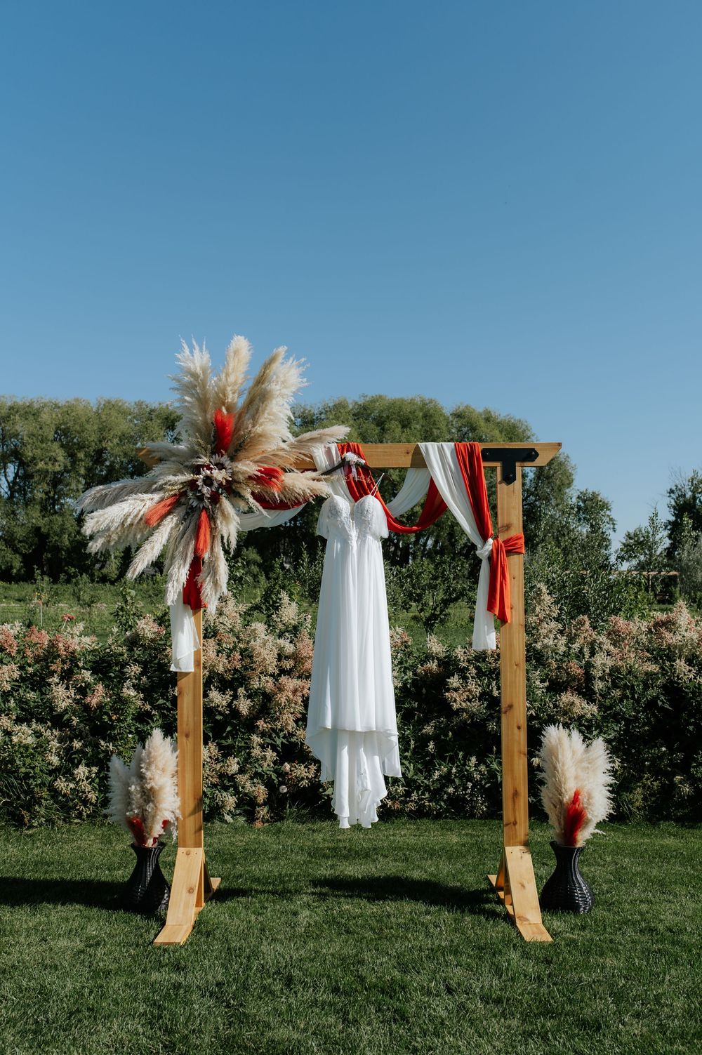 Rustic wooden wedding arch adorned with pampas grass, red ribbons, and white draping on a green lawn.