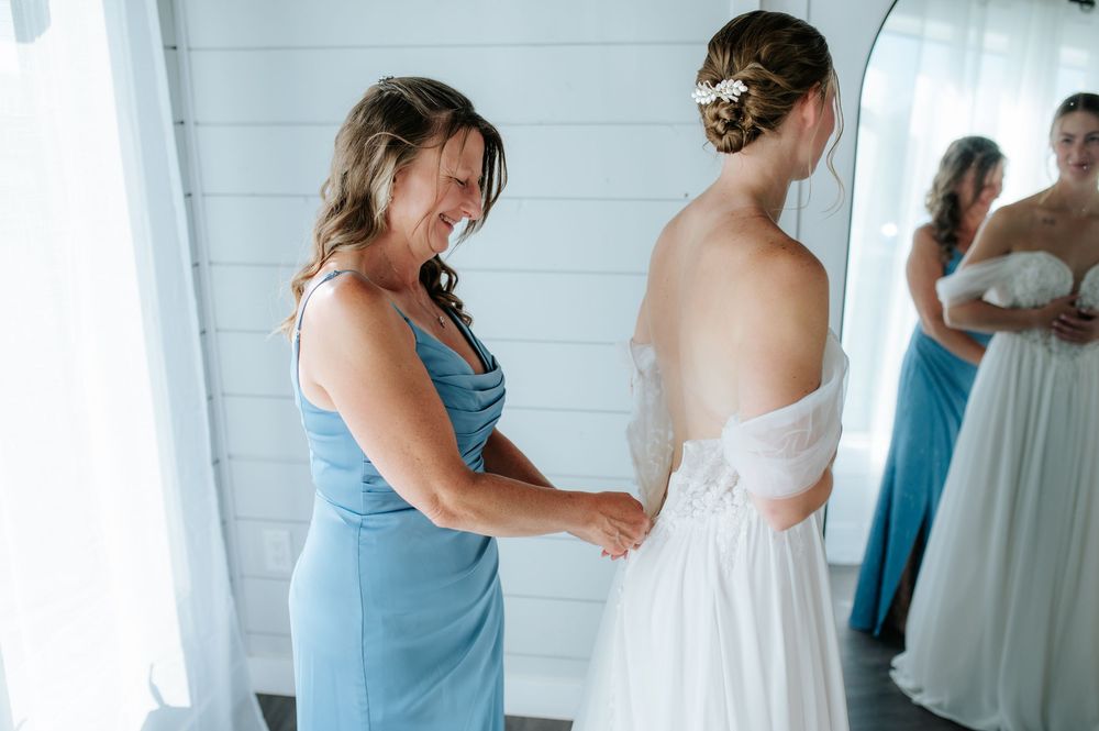 Bridesmaid in blue dress helping bride with wedding gown while another bridesmaid watches in mirror.