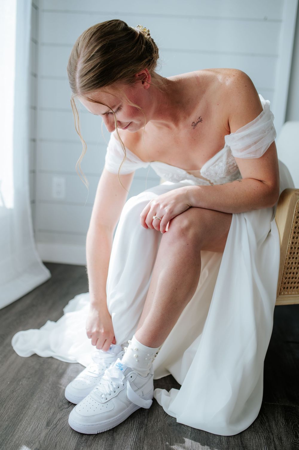Bride in an off-shoulder white wedding gown putting on white Nike Air Force 1 sneakers with lace socks while seated indoors.