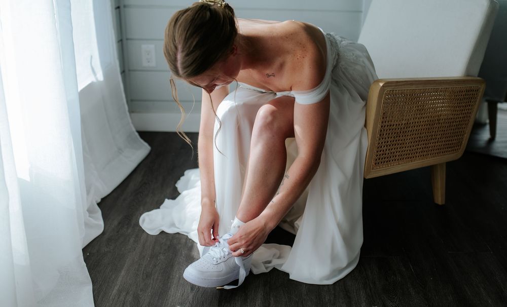 Bride in white off-shoulder wedding dress sitting in chair tying white sneakers on wedding day.