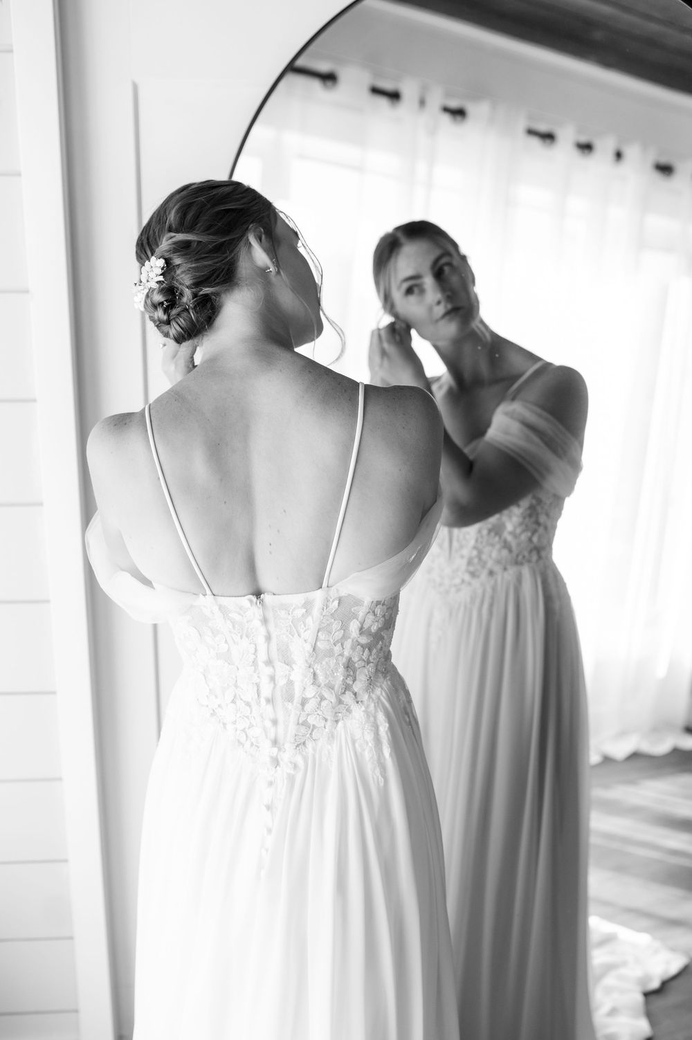 Black and white photo of bride in lace wedding gown adjusting earring while looking in round mirror.