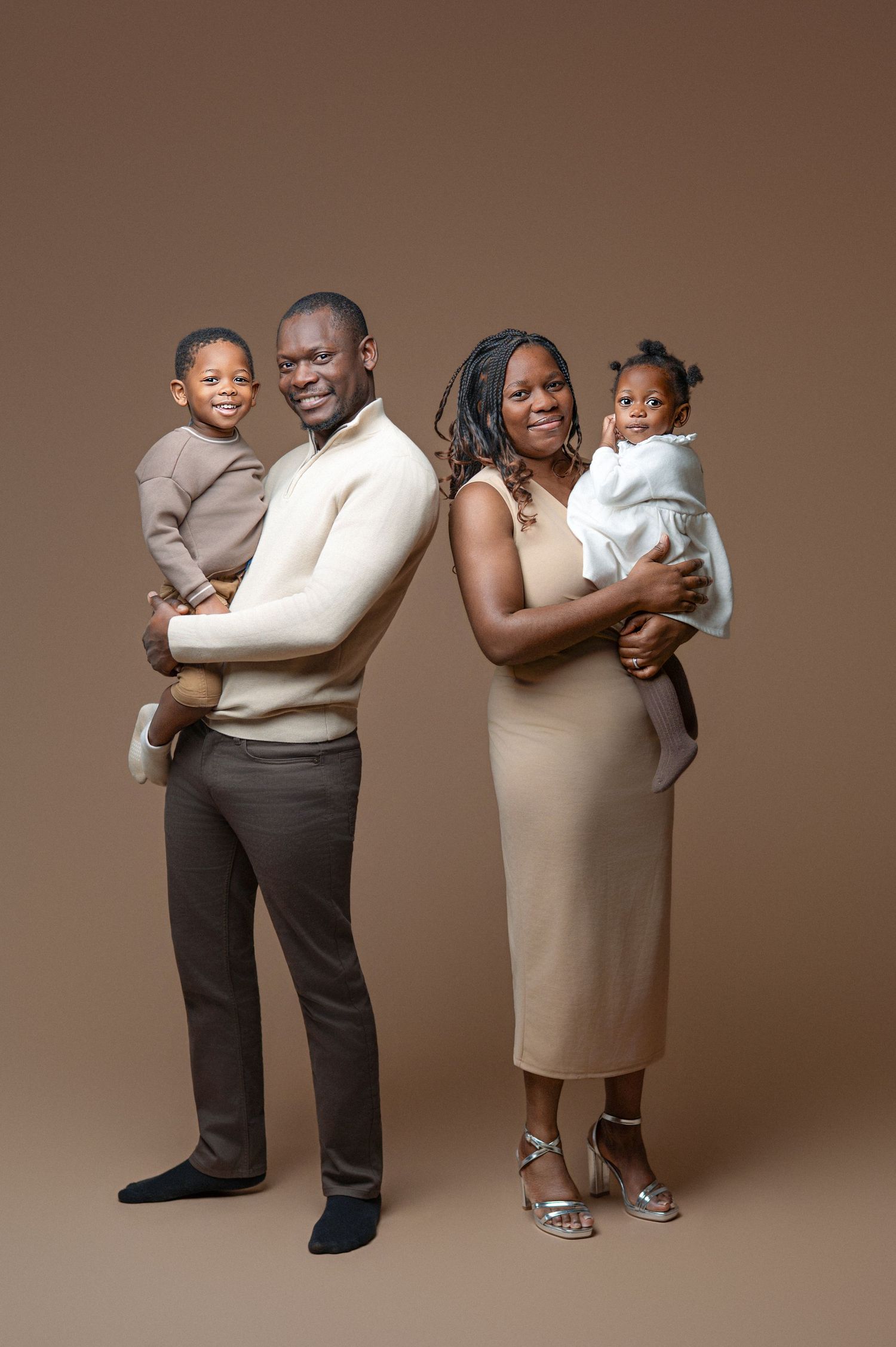 A smiling family of three poses in a studio, with parents holding their happy toddler against a gray background.