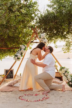 Couple sharing a kiss during a romantic beach proposal under a floral teepee arch with rose petals on sand.