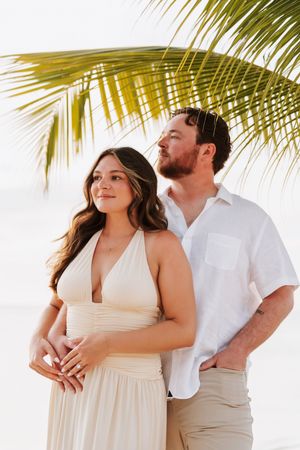 Couple posing under palm tree on beach, woman in cream halter dress, man in white linen shirt, tropical setting.