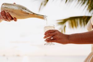 Person pouring Bottega prosecco into a glass outdoors near a palm tree at a tropical wedding celebration.