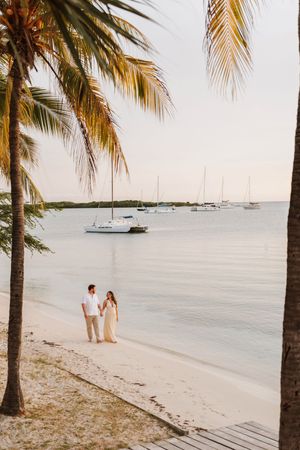 Couple holding hands on tropical beach at sunset with palm trees and sailboats on calm water.
