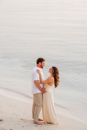 Couple embracing on a sandy beach at sunset, woman in cream gown and man in white shirt with tan pants.