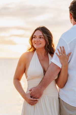 A couple shares a romantic beach sunset moment, woman in white halter dress smiling warmly at her partner.