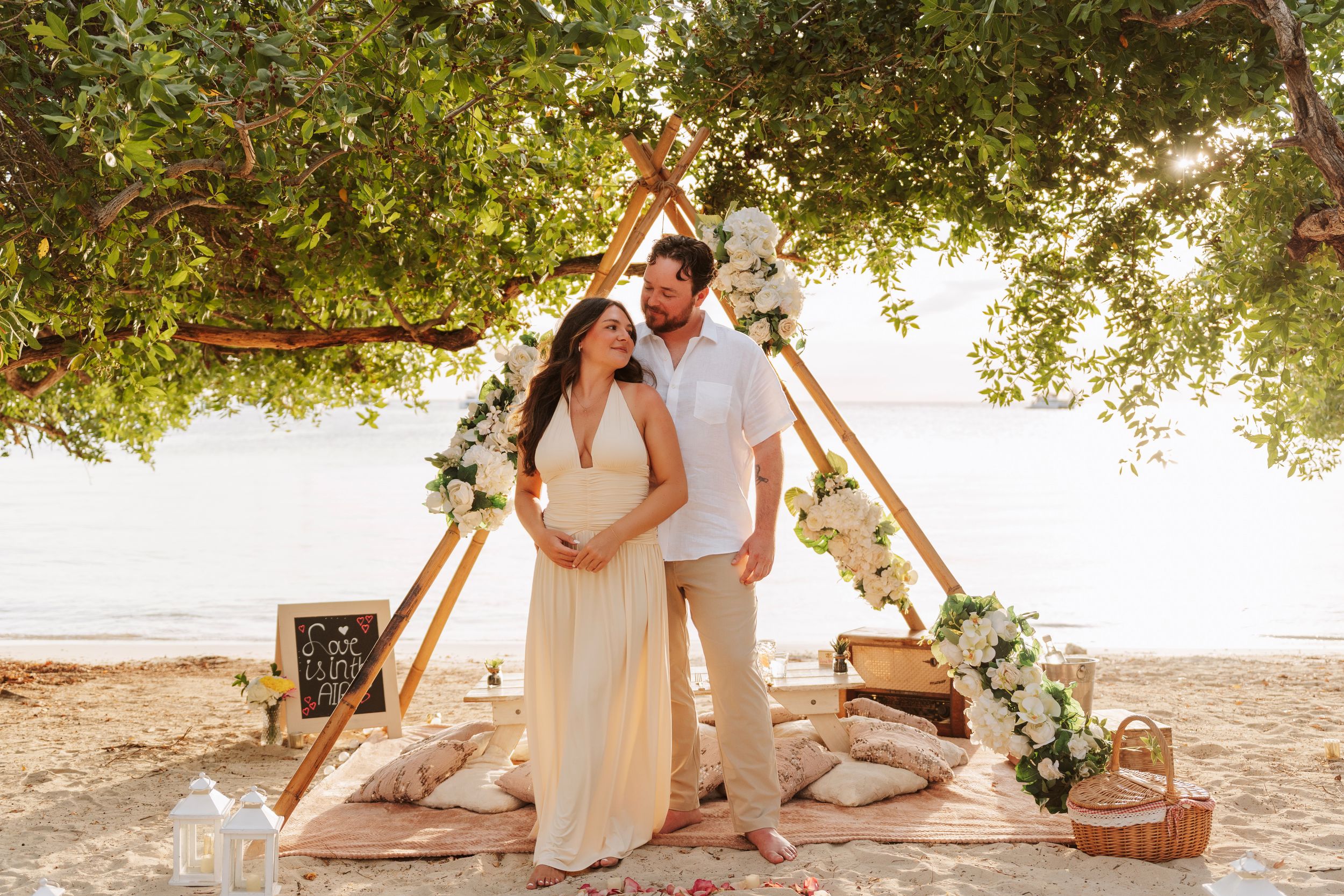 Couple poses under a wooden arch with flowers on a sandy beach at sunset during a romantic outdoor ceremony.
