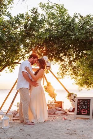 Couple embracing at sunset beach engagement setup with 'She Said Yes!' chalkboard sign under a tree.