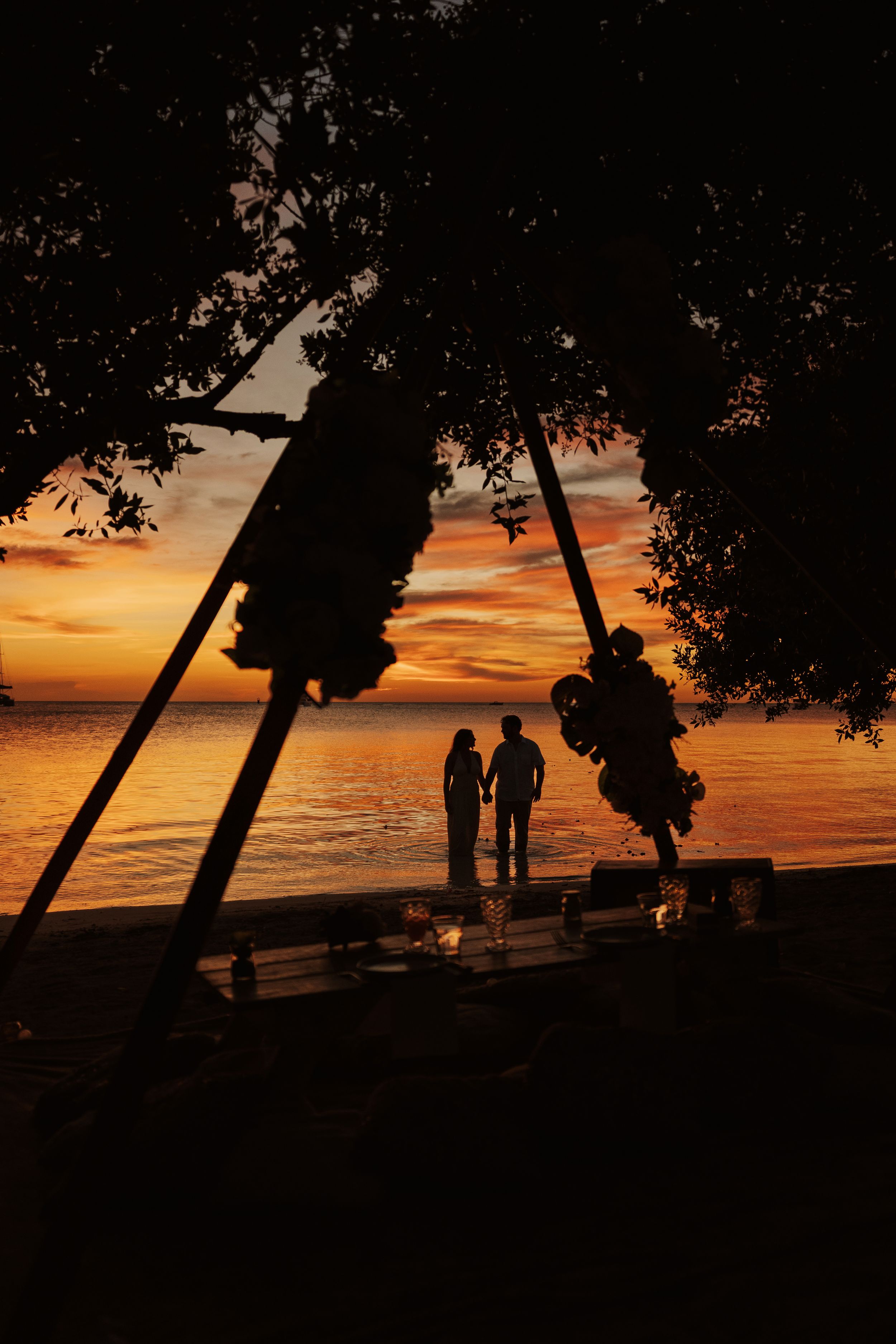 Silhouette of a couple holding hands on a tropical beach at sunset, framed by leaning palm trees.