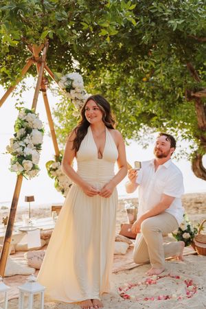 Man proposes to woman in white dress on sandy beach under tree with floral arch and rose petals.