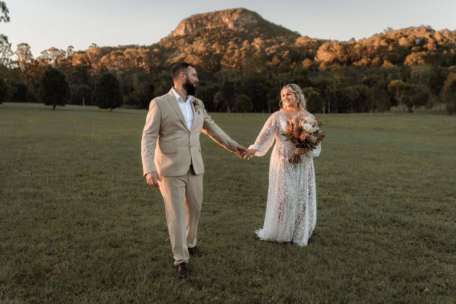 Bride and groom holding hands in a green field at golden hour with a mountain backdrop on their wedding day.