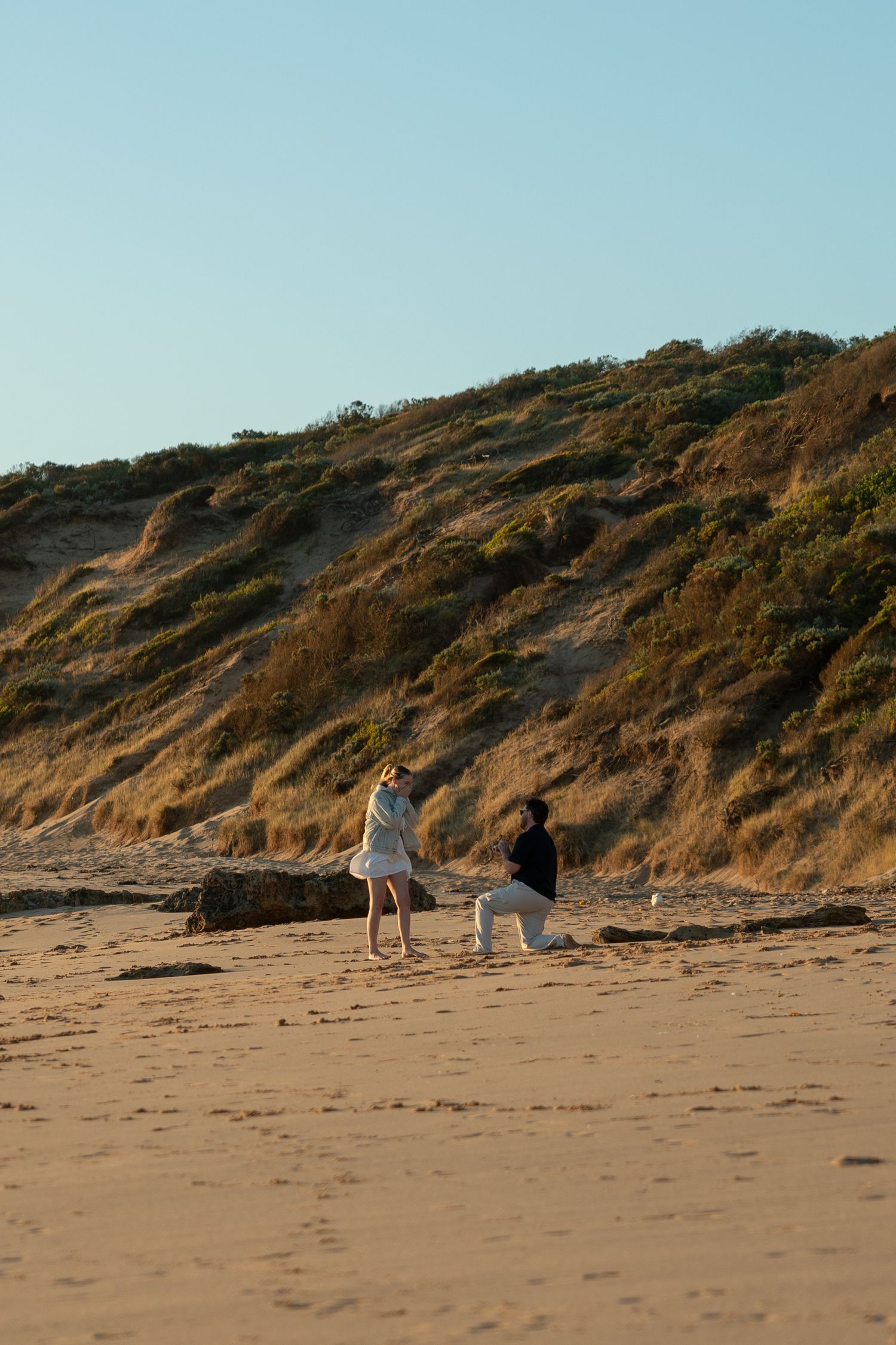 A couple shares a romantic beach proposal moment on sandy shores with golden dunes and clear blue sky.