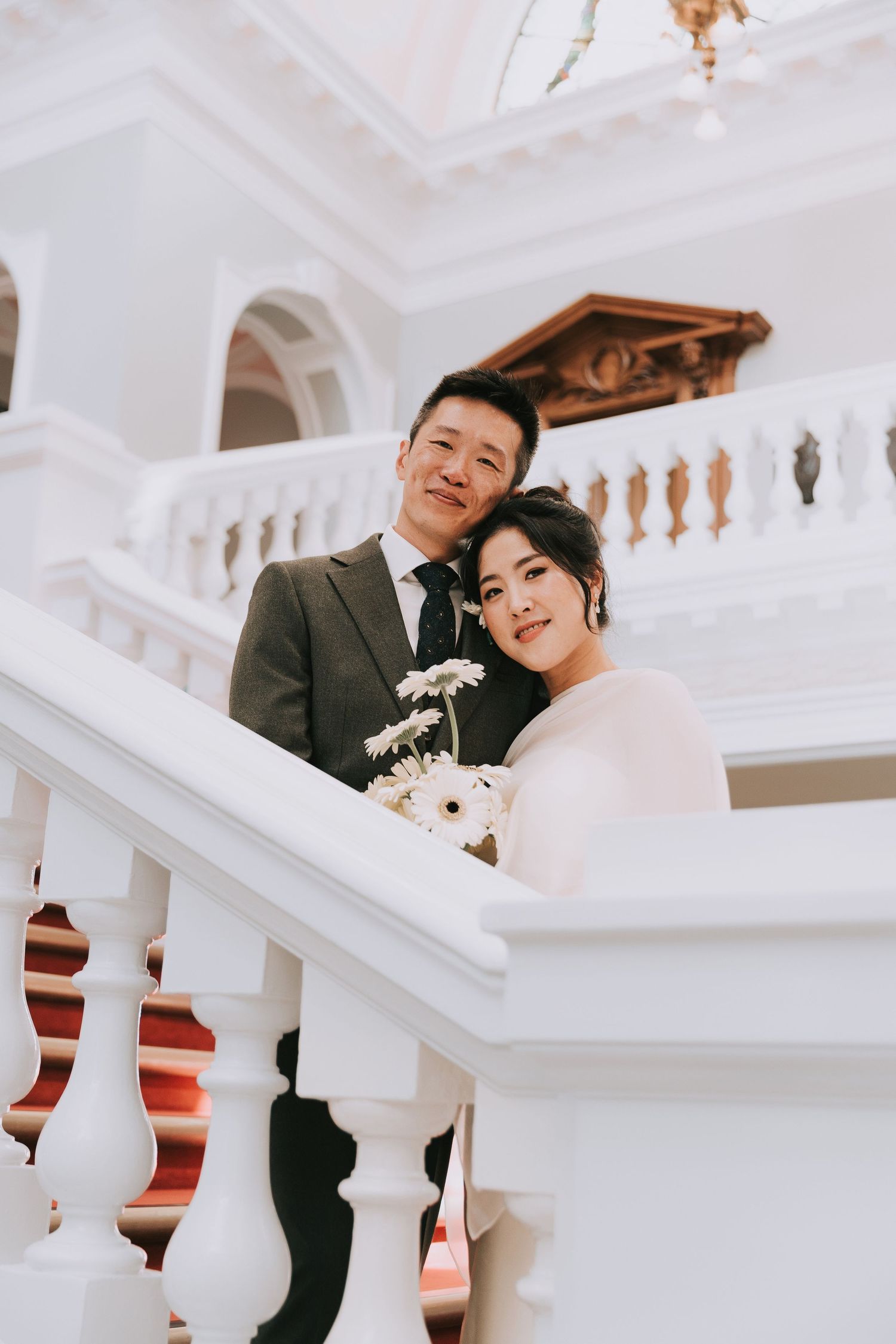 Bride and groom in wedding attire poses on on Grand Marble Staircase at Woolwich Town Hall , the bride holding white gerbera daisies.