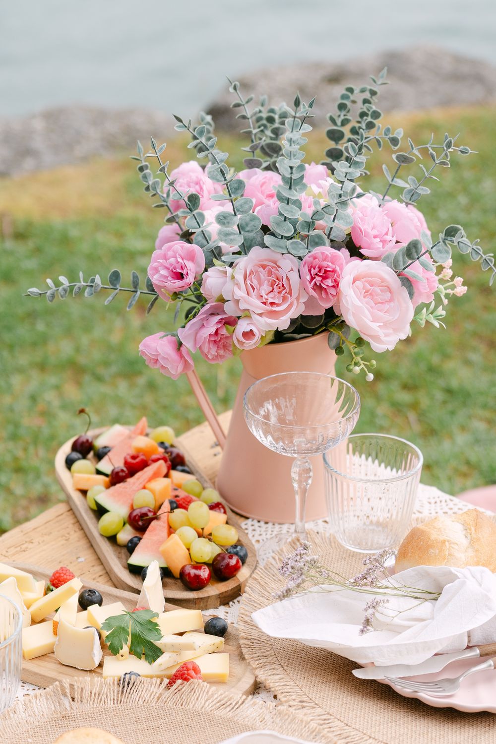 Elegant outdoor picnic table with pink roses, crystal glasses, cheese board, and fresh fruit display.