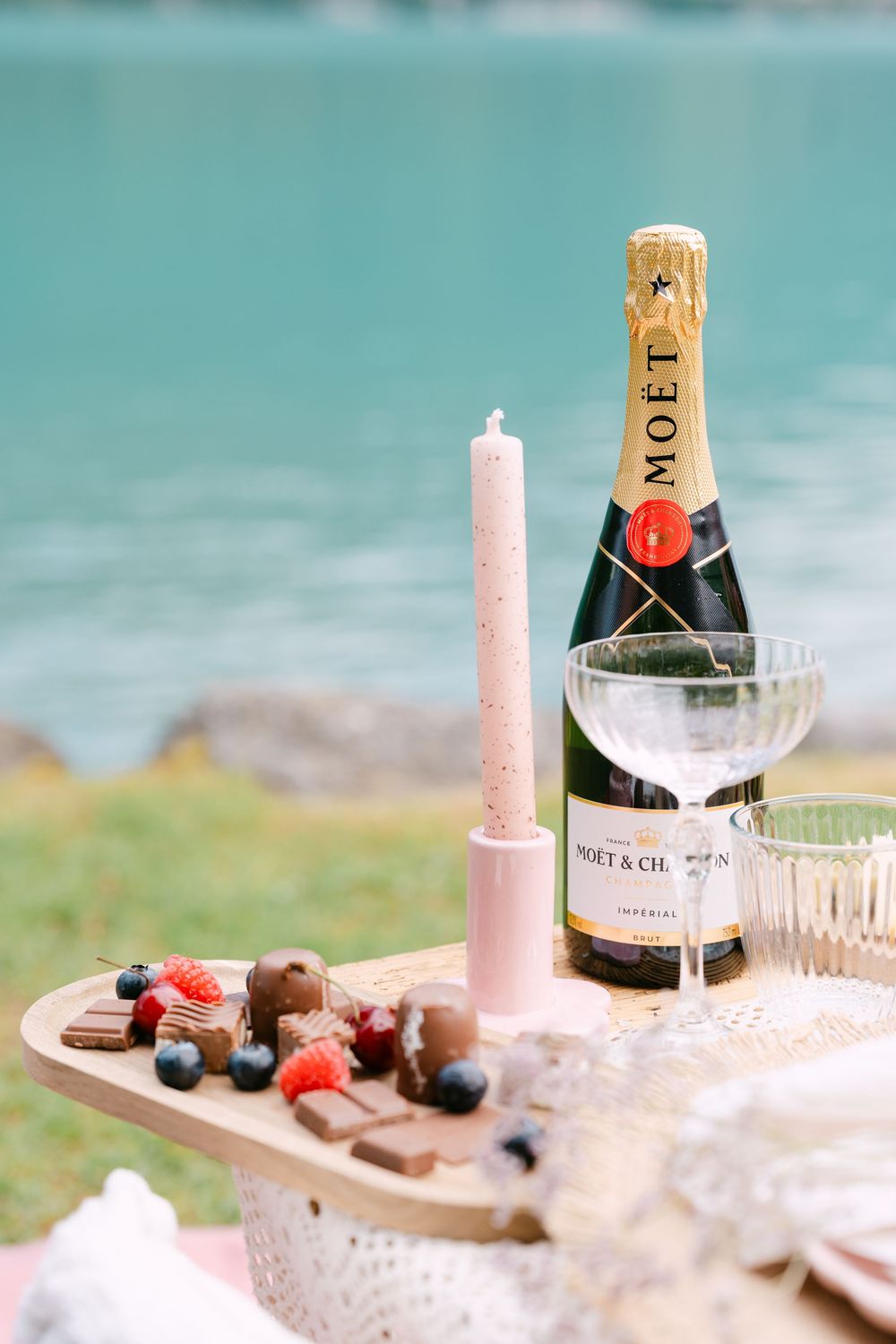 Moët & Chandon champagne bottle with coupe glasses, pink candle, and chocolate berry charcuterie board by turquoise lake.