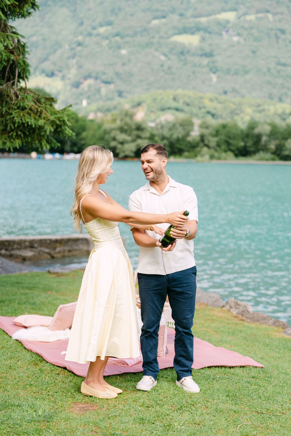 Couple celebrating with champagne on a lakeside picnic blanket, surrounded by lush greenery and turquoise water.