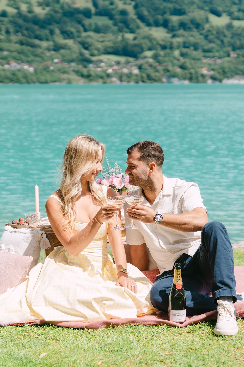 Couple toasting champagne glasses at a romantic lakeside picnic on a sunny day with turquoise water in the background.