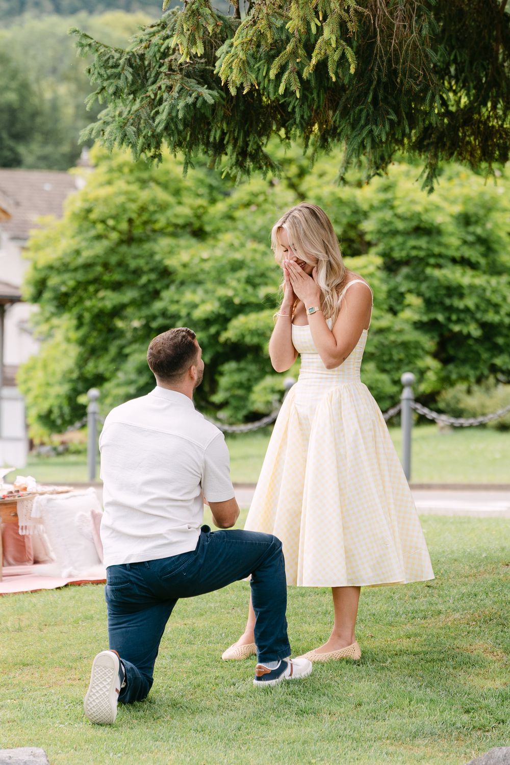 Man kneeling on one knee proposing to a surprised woman in a yellow dress outdoors on a green lawn.
