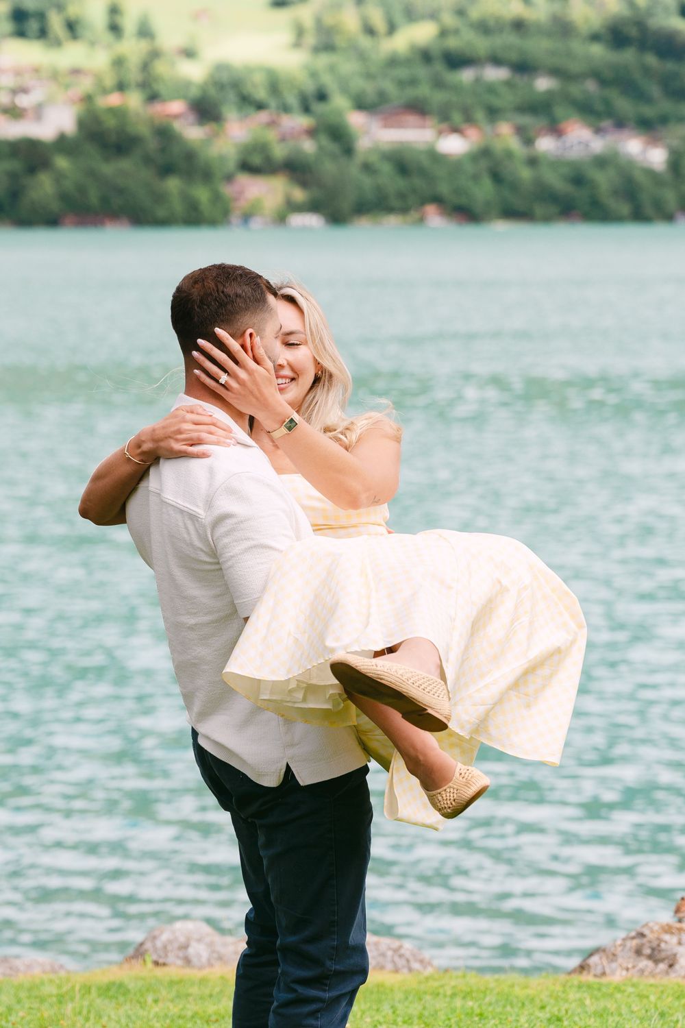 Man lifting woman in yellow dress by a turquoise lake, sharing a romantic moment with scenic hills in background.