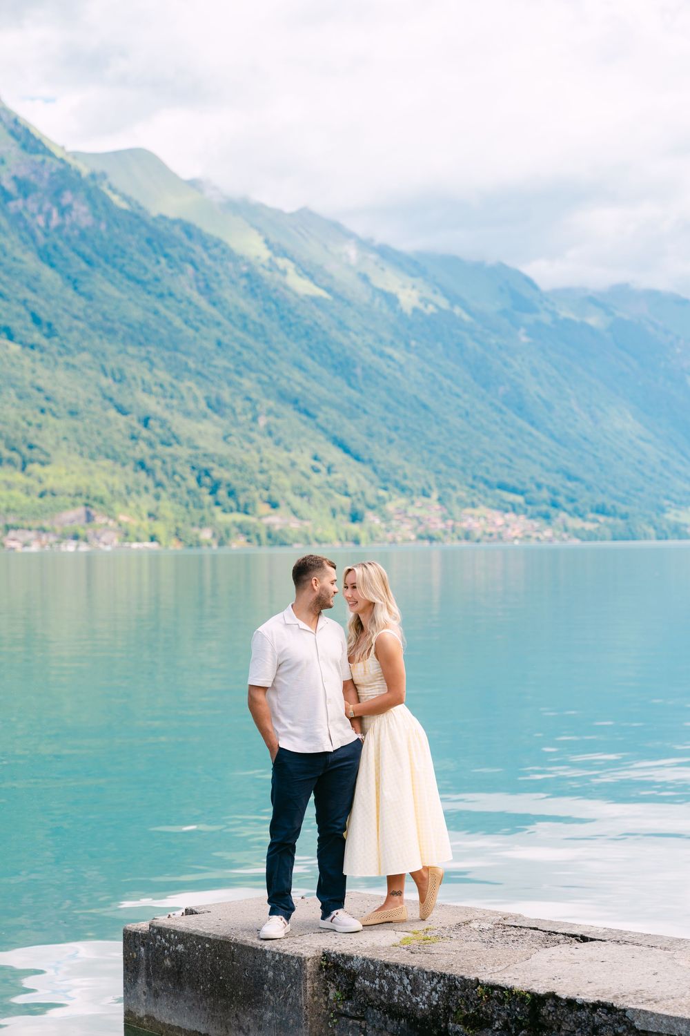 Romantic couple embracing on a stone pier with turquoise alpine lake and green mountains in background.