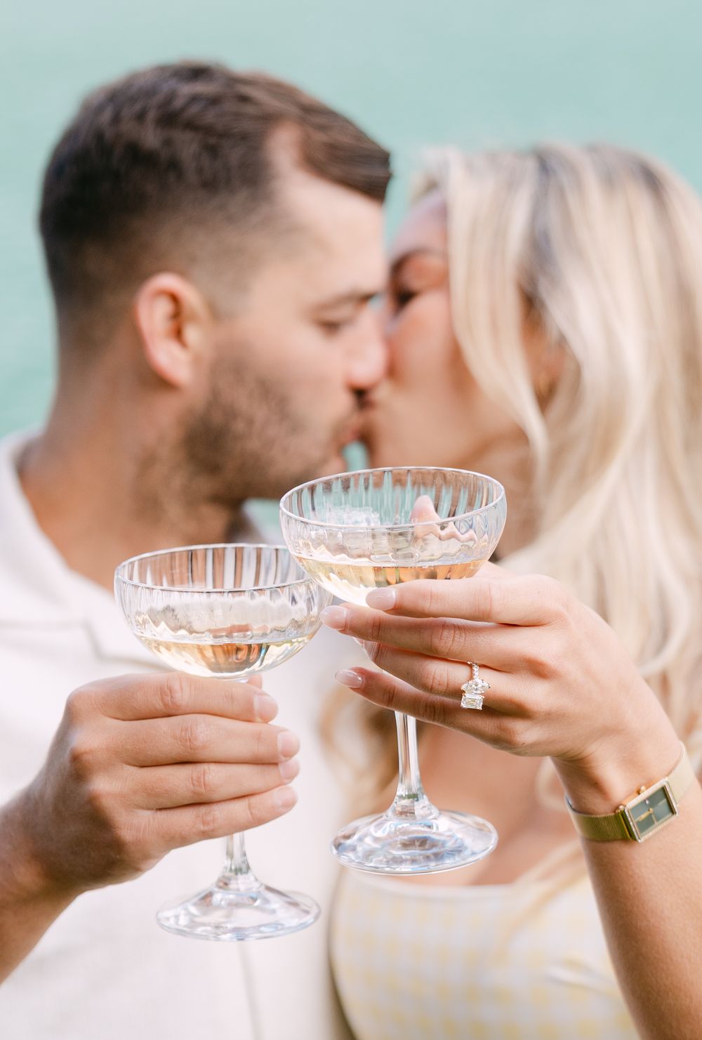 Engaged couple kissing while clinking crystal champagne coupe glasses, diamond engagement ring visible on woman's hand.