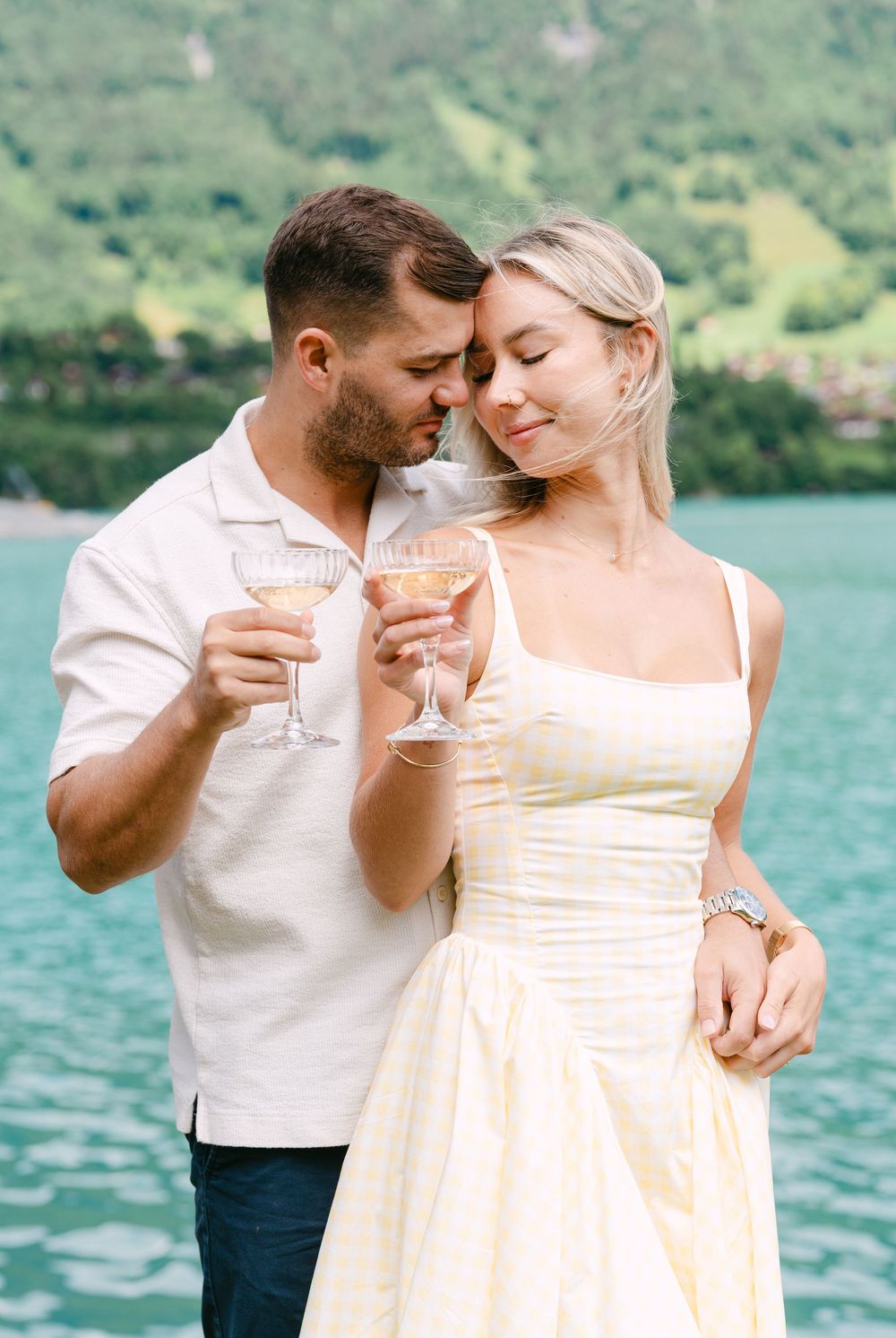 Couple toasting with champagne glasses by a turquoise lake, woman in yellow dress, man in white shirt, romantic moment.