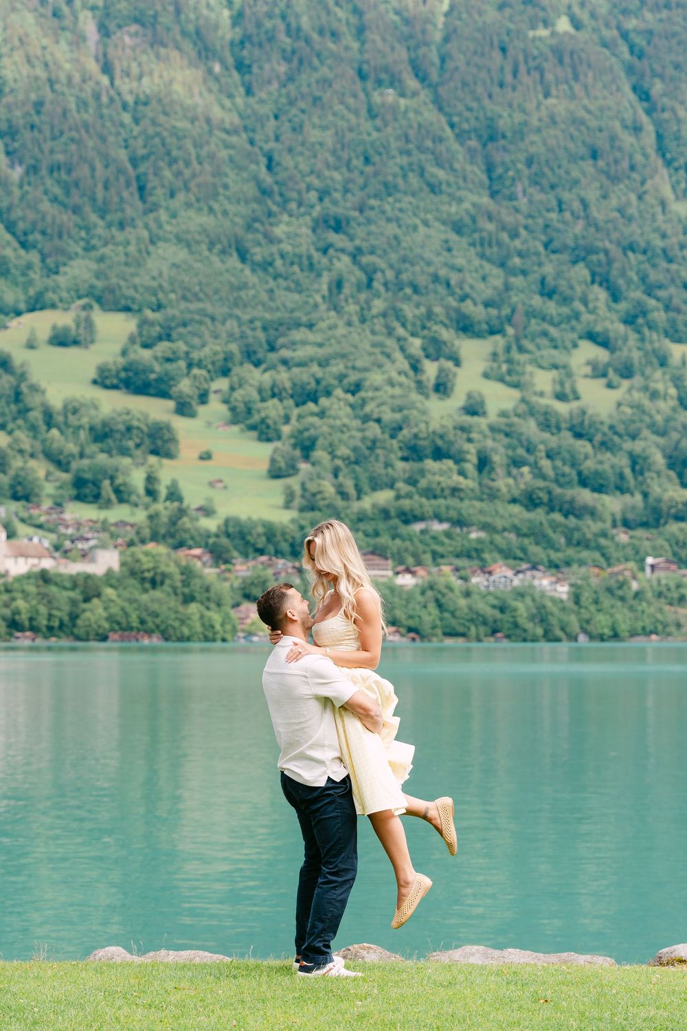 Man lifting woman by turquoise alpine lake with lush green mountain backdrop on sunny day.