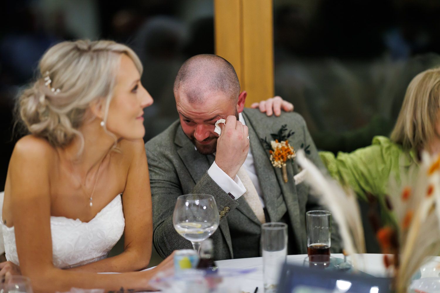 Emotional groom wiping tears at wedding reception while bride comforts him with hand on shoulder at decorated table.