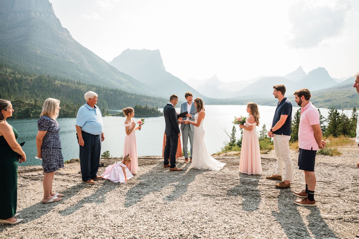Intimate outdoor wedding ceremony by a mountain lake with guests in formal attire and stunning rocky peaks backdrop.