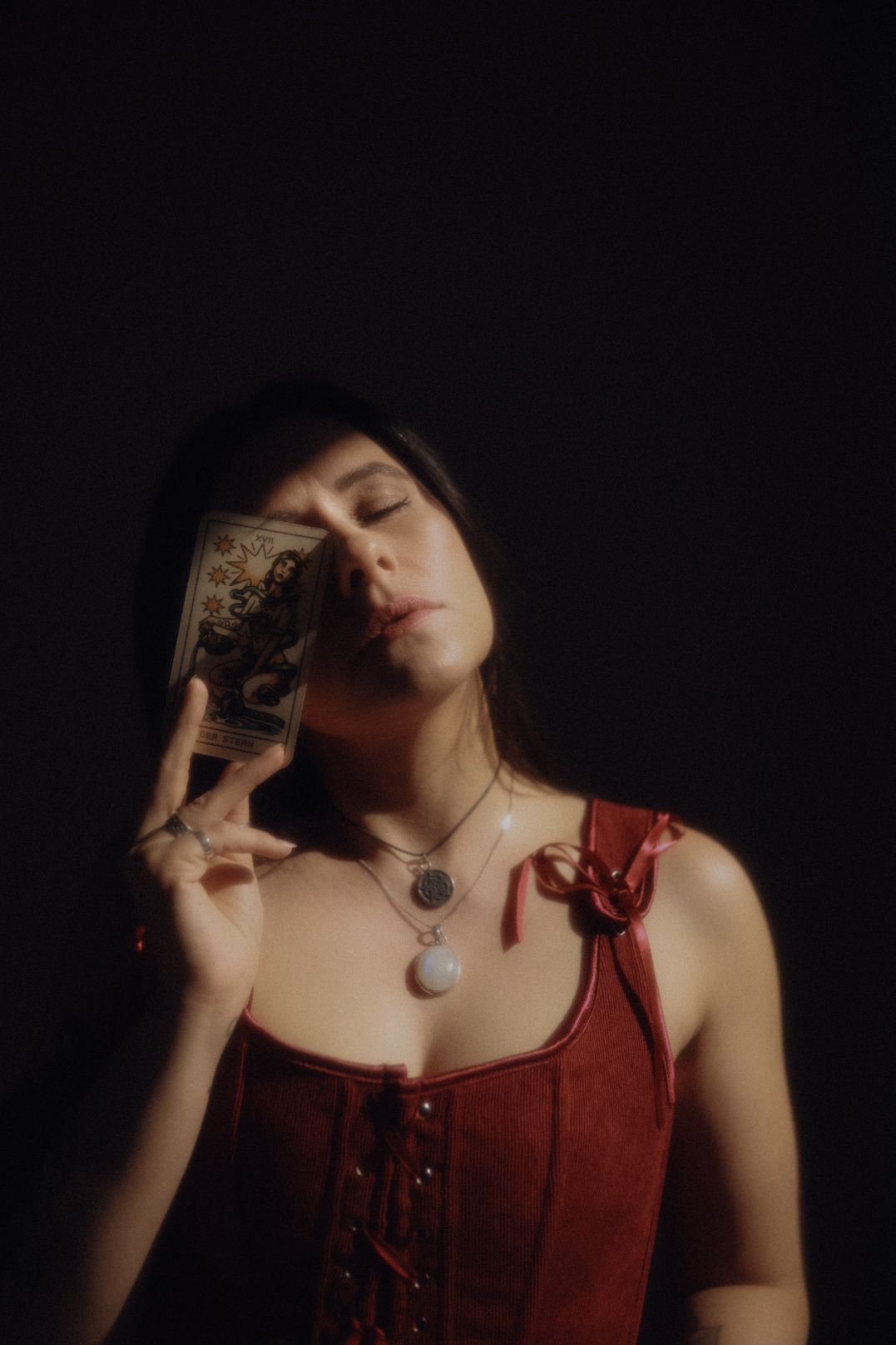 Woman in red corset holding tarot card with eyes closed, wearing layered necklaces against dark background.